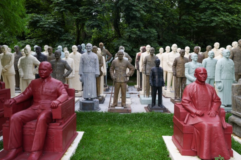 Statues of former Taiwan leader Chiang Kai-shek lined up in a park. Two of the statues in front show him seated. They are painted red. Some behind are standing. They are white or bronze.