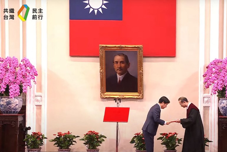 William Lai Ching-te during his inauguration ceremony. There is a flag of Taiwan on the wall and a portrait of Sun Yat-sen, founder of the Republic of China. with large displays of pink orchids on either side. Lai is bowiing as he receives a document from an official.