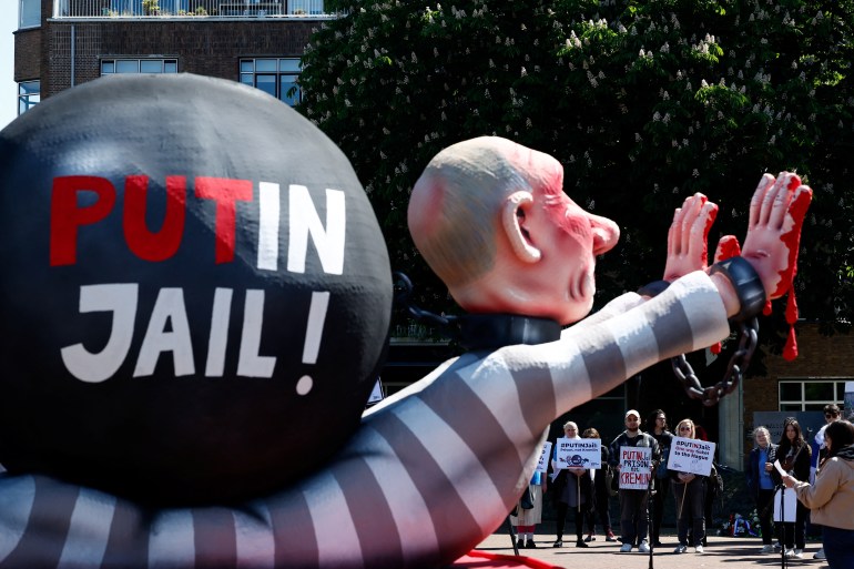 A giant effigy of Vladimir Putin in striped prison garb. Its hands are chained and painted red. 'Putin Jail' is written on a black ball on the puppet's back. Protesters are gathered nearby. They are holding placards calling for Putin to be jailed.