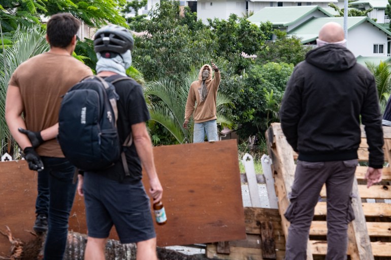 People facing each other at a barricade in Noumea. There are three men with their backs to the camera with another man standing on the other side. The man is masked and wearing a hoodie. He is gesturing towards them.