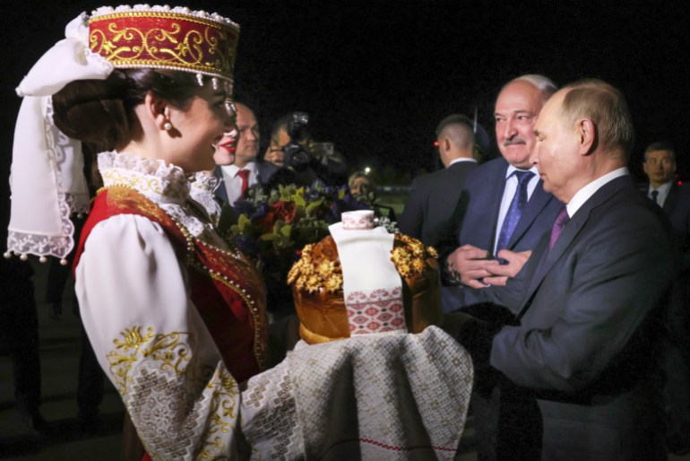 Alexander Lukashenko welcoming Vladimir Putin to Belarus. There s a woman in traditional dress on the left holding a gift. A woman behind her has a bouquet of flowers. Lukashenko and Putin are on the right.