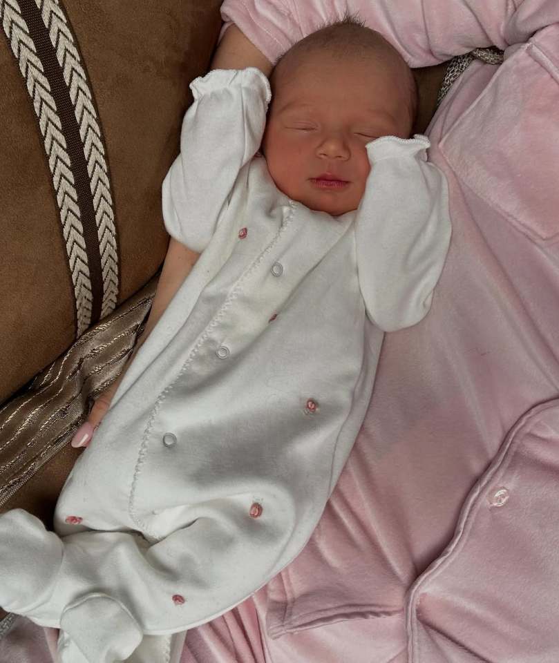 Sleeping newborn baby girl in white onesie held by a person wearing pink.