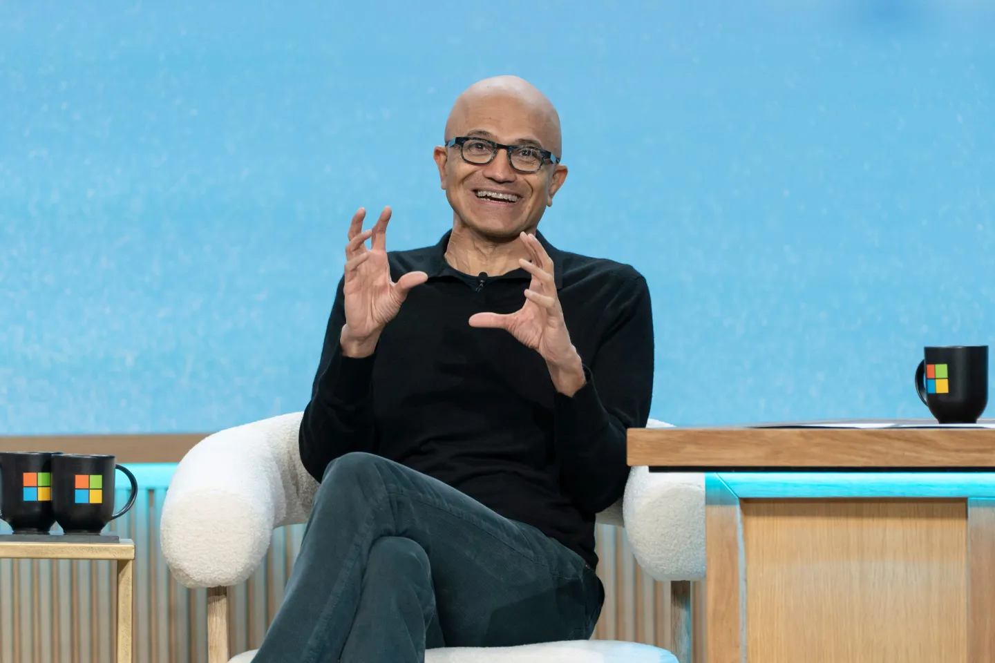 Microsoft CEO Satya Nadella sitting on stage at the company's 50th Anniversary celebrations, wearing a black long-sleeve shirt and black jeans, making a "globe"-like gesture with his two hands.